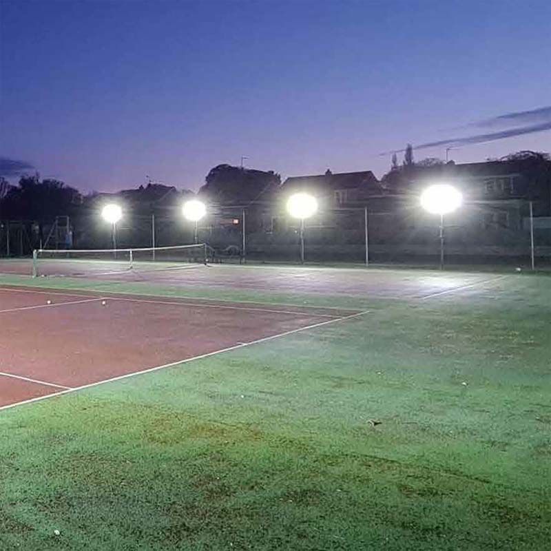 A tennis court illuminated at dusk by Ritelite Sports Lite Fence Fixed Floodlights, with houses and trees visible in the background beneath a darkening sky.