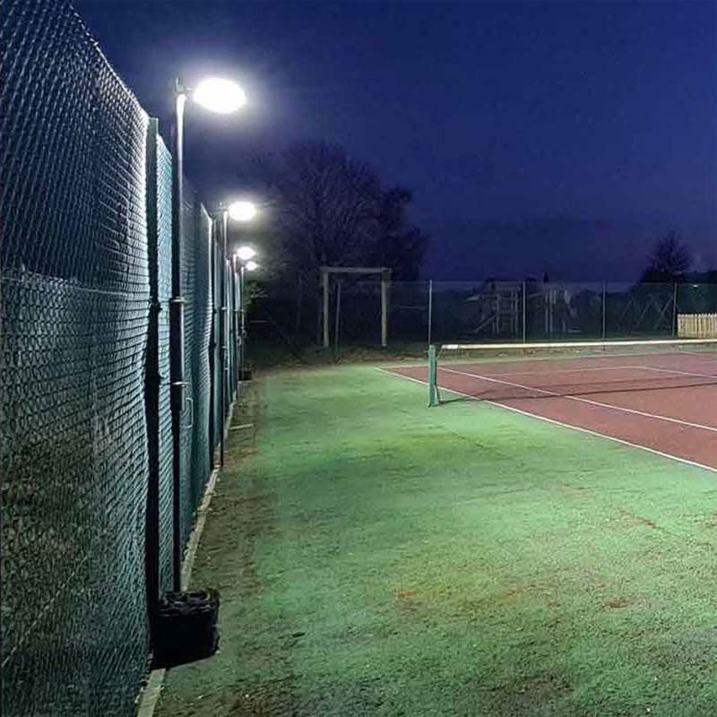 A tennis court at night is brightly lit by Ritelite Sports Lite Fence Fixed Floodlights, casting light over the green and red court surface while the surrounding area remains dark with trees visible in the background.