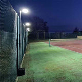 A tennis court at night is lit by Ritelite Sports Lite Fence Fixed Floodlights, casting a glow over the green and red surface while trees and playground equipment fade into the darkness beyond.