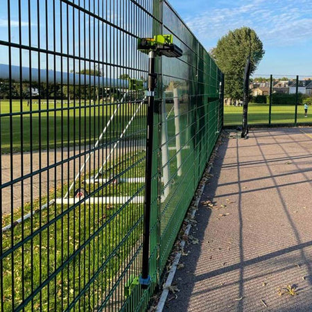 A camera on a tall tripod is attached to a green fence by a sports field near Ritelite Sports Lite Fence Fixed Floodlights, capturing the sunny scene with blue sky, clouds, and houses and trees in the background.