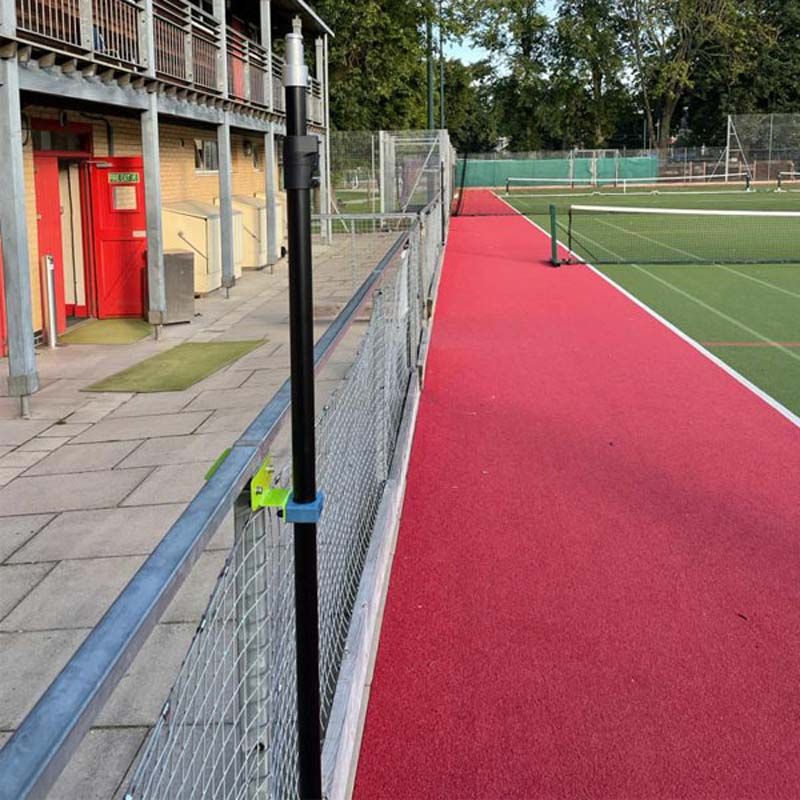 A red running track curves past a fenced sports field and a building with a balcony. Ritelite Sports Lite Fence Fixed Floodlights are attached to the fence near a black pole, with trees visible in the background.