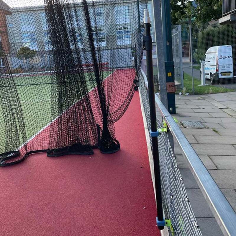 A torn net hangs over a red sports court, partially blocking the entrance. Ritelite Sports Lite Fence Fixed Floodlights line the fence, with a sidewalk and white van visible outside the perimeter.