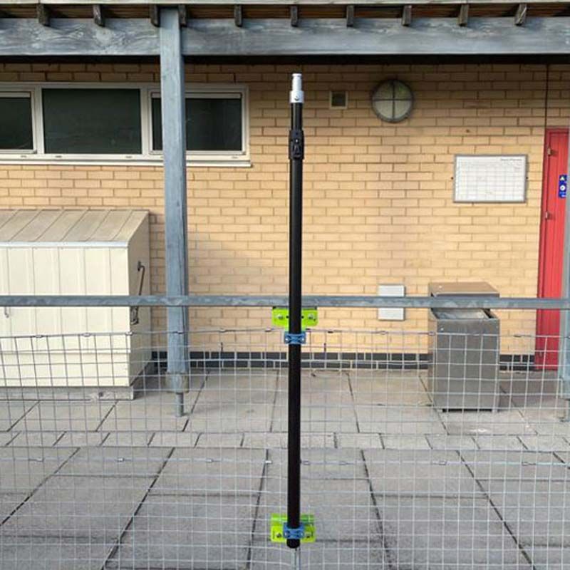 A Ritelite Sports Lite Fence Fixed Floodlight is mounted on a black vertical pole with green brackets to a wire fence in front of a brick building with windows, red door, and utility boxes—perfect for reliable sports area lighting.