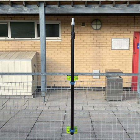 A Ritelite Sports Lite Fence Fixed Floodlight is mounted on a black vertical pole with green brackets to a wire fence in front of a brick building with windows, red door, and utility boxes—perfect for reliable sports area lighting.