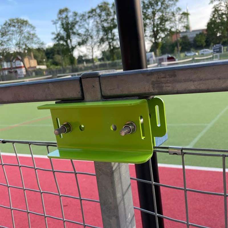 A close-up of a Ritelite Sports Lite Fence Fixed Floodlights unit, attached to a metal railing with a green bracket at a sports field, with battery powered lights illuminating the grass, track, and nearby trees.