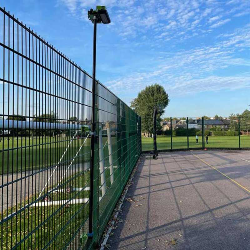 A tall metal fence with Ritelite Sports Lite Fence Fixed Floodlights separates the sports court from a grassy field. Fence-mounted lights illuminate the area, while trees and houses appear in the distance under a blue, cloud-scattered sky.