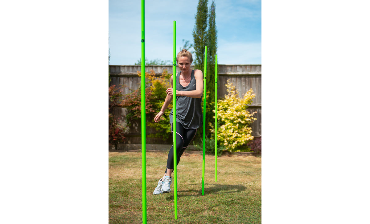 A woman in athletic clothing skillfully weaves through the Finesse Agility Pole - Set of 6 by Finesse, set up on a sunny grassy lawn as she focuses on her outdoor exercise routine.