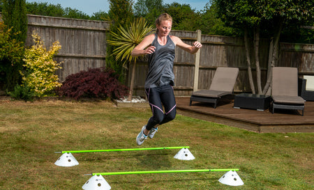 A woman in athletic wear jumps over Finesse Agility Poles (Set of 6) arranged as an outdoor obstacle course on a grassy lawn, with patio chairs and plants in the background.