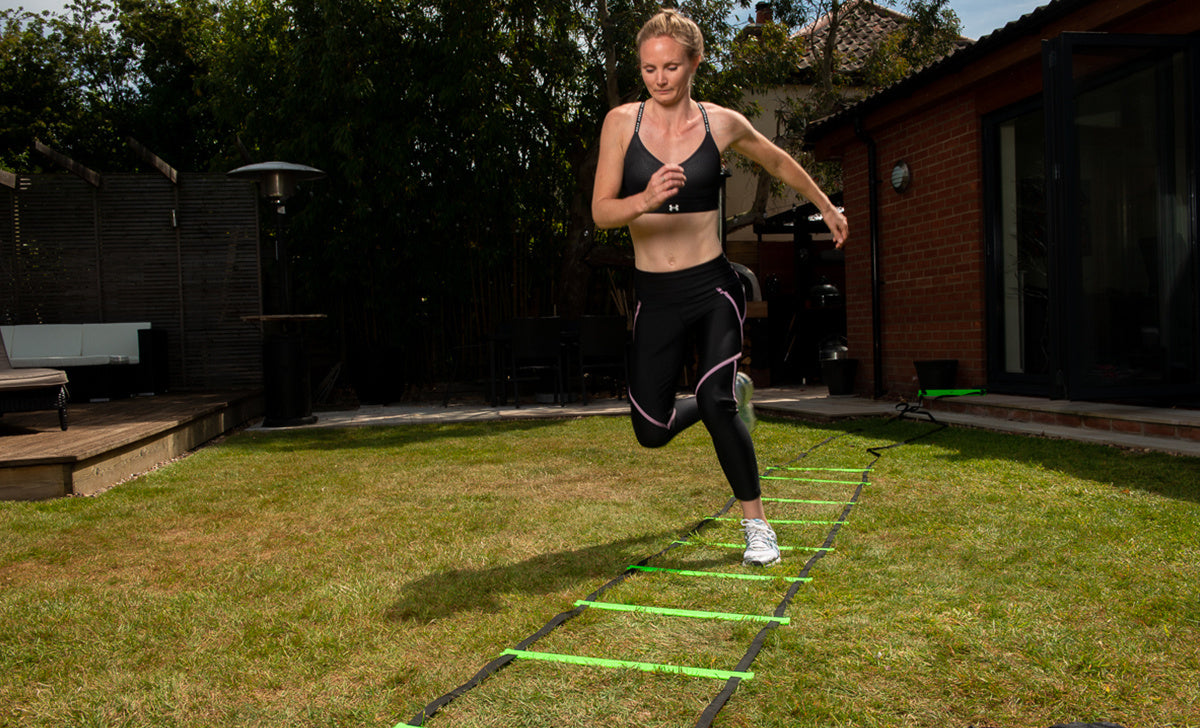 A woman in athletic clothing uses the Finesse 4m Speed Ladder by Finesse, doing agility ladder drills on a grassy lawn outside, focused and mid-step, with a house and trees visible in the background.