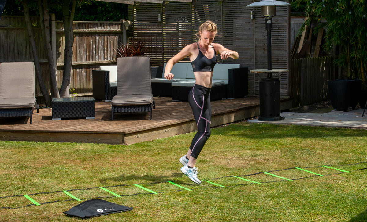 A woman in athletic wear trains on grass in her backyard using the Finesse 4m Speed Ladder by Finesse, surrounded by patio furniture and greenery.