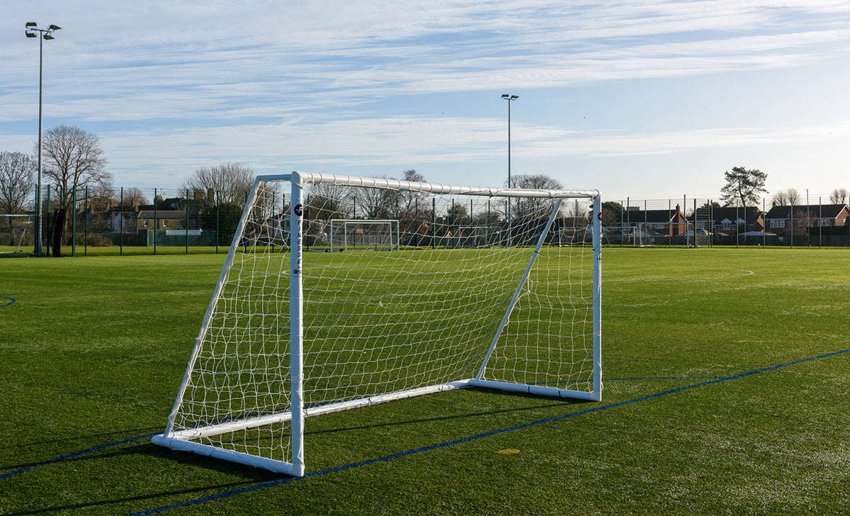 A Finesse 12 x 6ft Match Goal by Finesse stands on green artificial turf beneath a blue sky with light clouds. Trees, houses, and another match goal appear in the background.