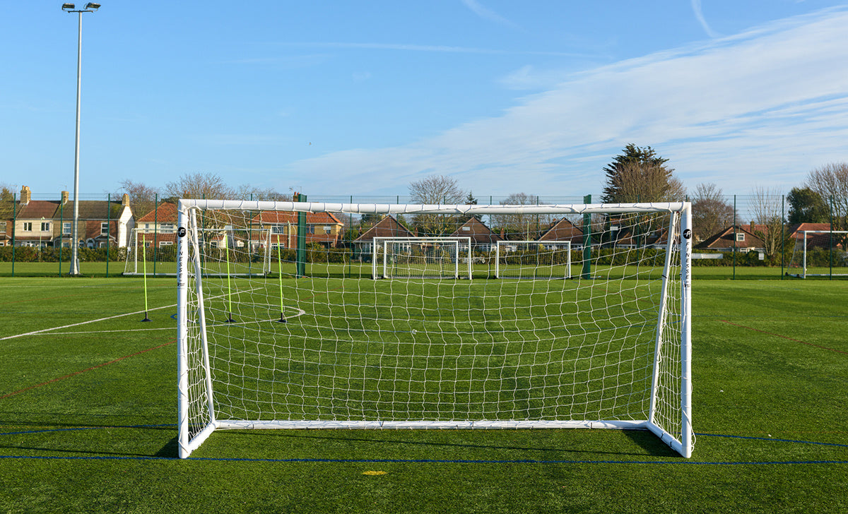 A soccer field featuring several Finesse 12 x 6ft Match Goals by Finesse, aligned in a row on green artificial turf, with houses and trees visible in the background beneath a clear blue sky.