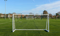 A soccer field featuring several Finesse 12 x 6ft Match Goals by Finesse, aligned in a row on green artificial turf, with houses and trees visible in the background beneath a clear blue sky.