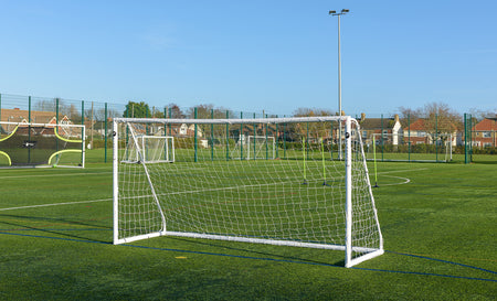 The Finesse 12 x 6ft Standard Goal by Finesse stands on green artificial turf, with other regulation goals, netted fencing, and houses in the background under clear skies.