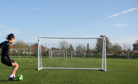 A soccer player in black sportswear gets ready to kick a green and black ball toward an empty Finesse 12 x 6ft Standard Goal on grass, with more Finesse goals and houses visible in the background.