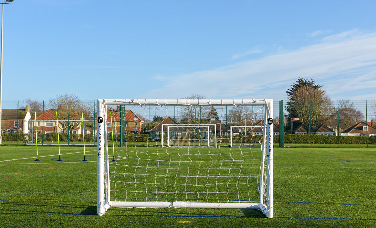 A Finesse 6 x 4ft Standard Goal by Finesse stands on green artificial turf under a clear blue sky, with additional goals and houses visible in the background.
