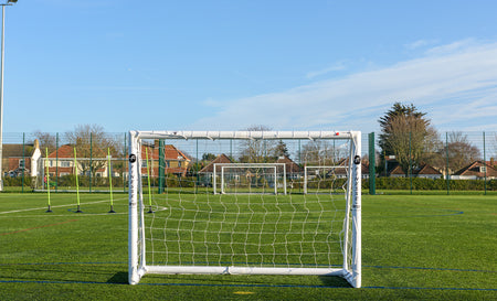 The Finesse 8 x 4ft Standard Goal by Finesse sits on green artificial turf, with other goals and netted fences nearby under a clear blue sky. Houses and trees surround the field, making it ideal for garden football games.