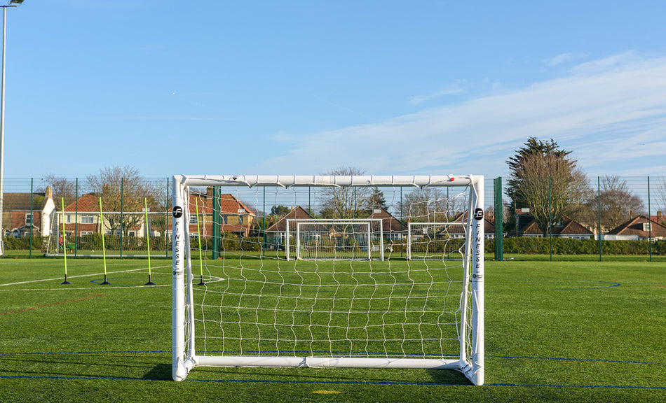 The Finesse 8 x 4ft Standard Goal by Finesse sits on green artificial turf, with other goals and netted fences nearby under a clear blue sky. Houses and trees surround the field, making it ideal for garden football games.