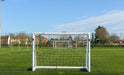 The Finesse 8 x 4ft Standard Goal by Finesse sits on green artificial turf, with other goals and netted fences nearby under a clear blue sky. Houses and trees surround the field, making it ideal for garden football games.