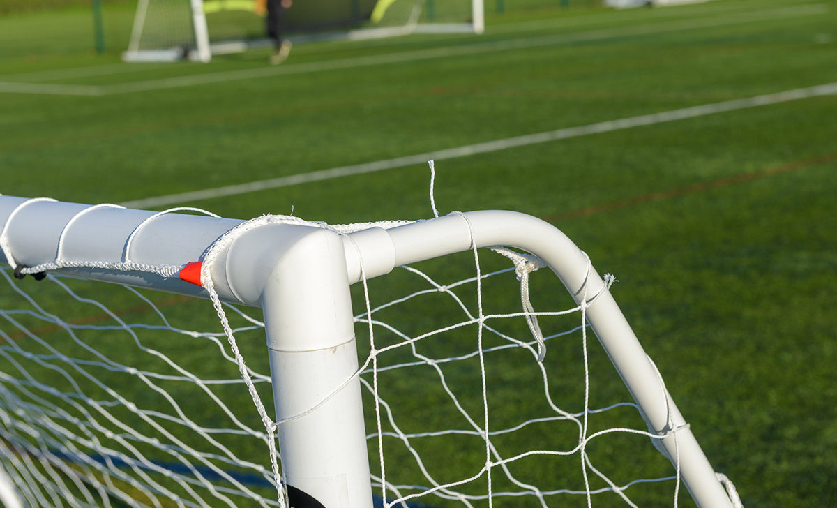 A close-up of the Finesse 6 x 4ft Standard Goal for kids by Finesse, featuring white netting on green artificial turf, with other goals and a blurred player in the background.