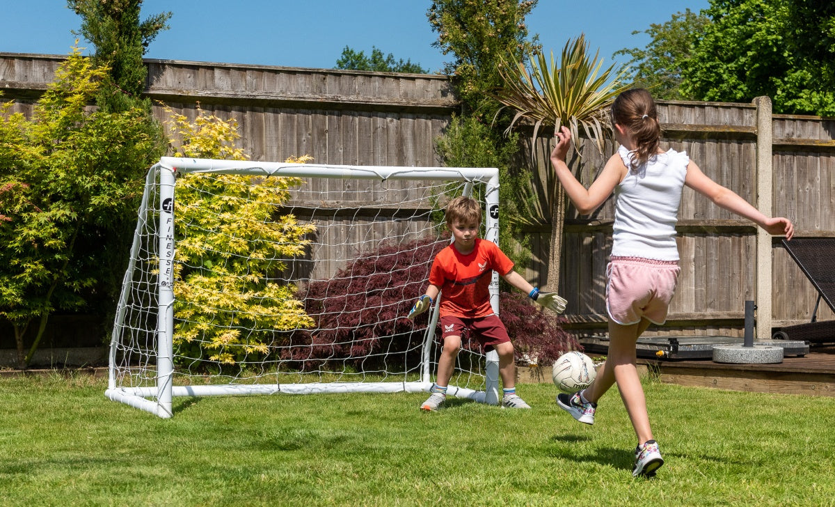A girl in white kicks a soccer ball toward the Finesse 8 x 4ft Standard Goal, while a boy in red stands ready as goalkeeper. They play on green grass under the sun, with plants and a wooden fence in the background.
