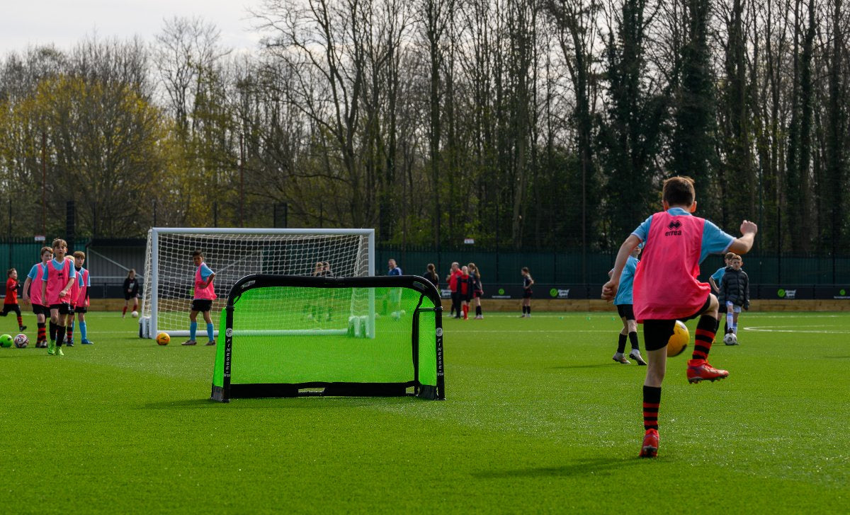 Children in sports bibs play soccer on a green field, aiming at the Finesse Folding Target Goal by Finesse. One kicks the ball as others watch or join in, with trees and a fence lining the background.