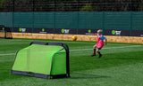 A young child in a pink training bib and hat kicks a soccer ball toward the Finesse Folding Target Goal by Finesse on an outdoor artificial turf field, with sports banners visible in the background under daylight.