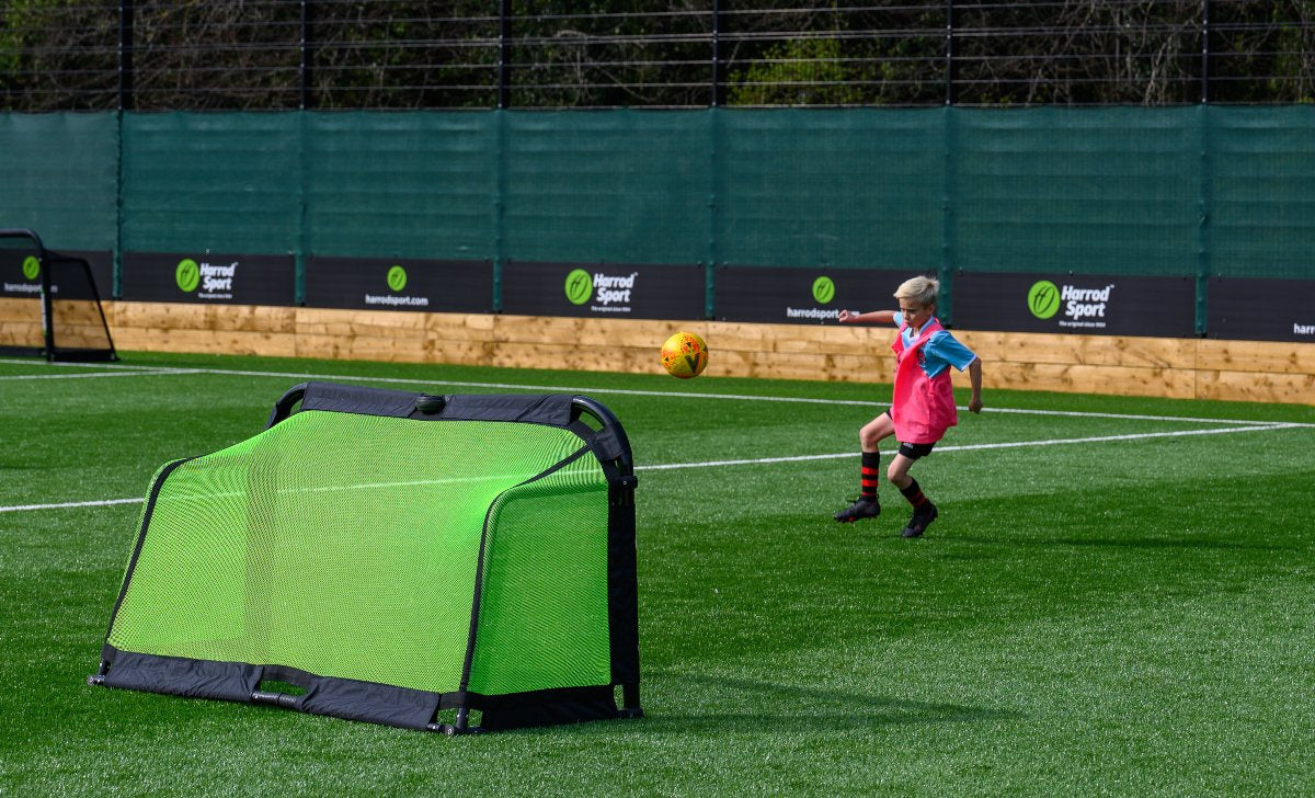 A young child in a pink training bib and hat kicks a soccer ball toward the Finesse Folding Target Goal by Finesse on an outdoor artificial turf field, with sports banners visible in the background under daylight.