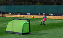A young child in a pink training bib and hat kicks a soccer ball toward the Finesse Folding Target Goal by Finesse on an outdoor artificial turf field, with sports banners visible in the background under daylight.
