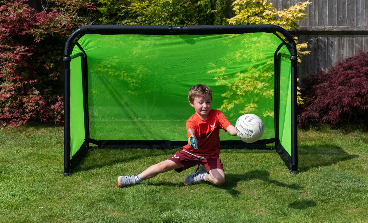 A young boy in sportswear kneels on grass in front of the Finesse Folding Target Goal by Finesse, catching a white soccer ball with one hand, with colorful bushes and a wooden fence in the background.