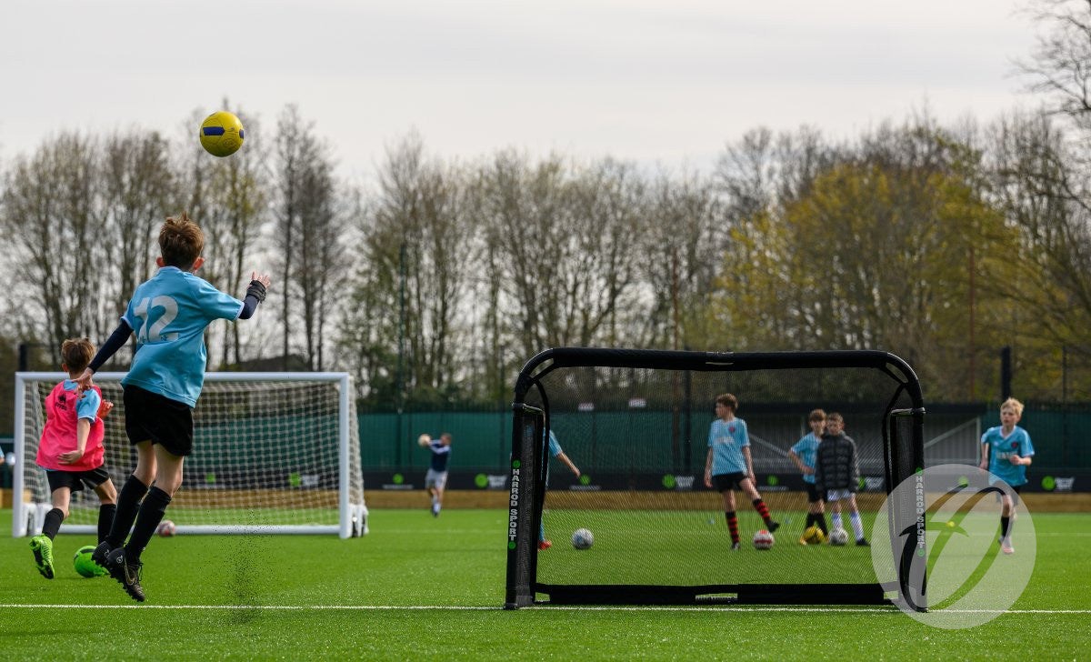 A youth soccer player in a blue jersey kicks a yellow ball towards the Finesse Folding Target Goal on a green outdoor field, with teammates and opponents watching and playing in the background.