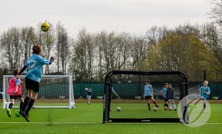 A youth soccer player in a blue jersey kicks a yellow ball towards the Finesse Folding Target Goal on a green outdoor field, with teammates and opponents watching and playing in the background.