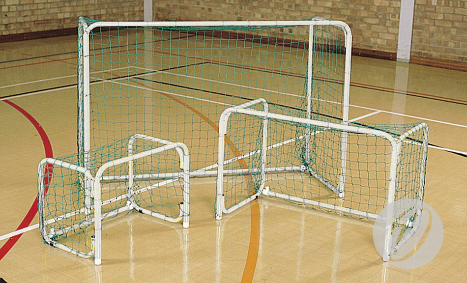 A Harrod Sport Mini Hoc Goal (1000MM x 1400MM) with a white folding frame and green net is set on an indoor gym floor marked with red and black lines, in front of a brick wall.