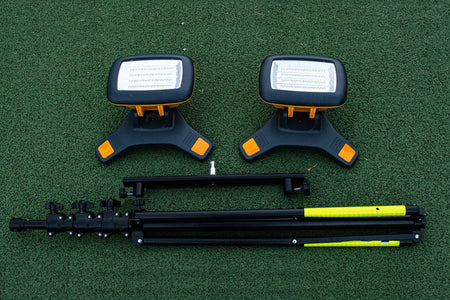 Two Nightsearcher Galaxy Pro Twin Portable Floodlights with orange accents and two folded black/yellow tripods are laid on a green surface, perfect as outdoor training lights or a rechargeable lighting kit for any activity.