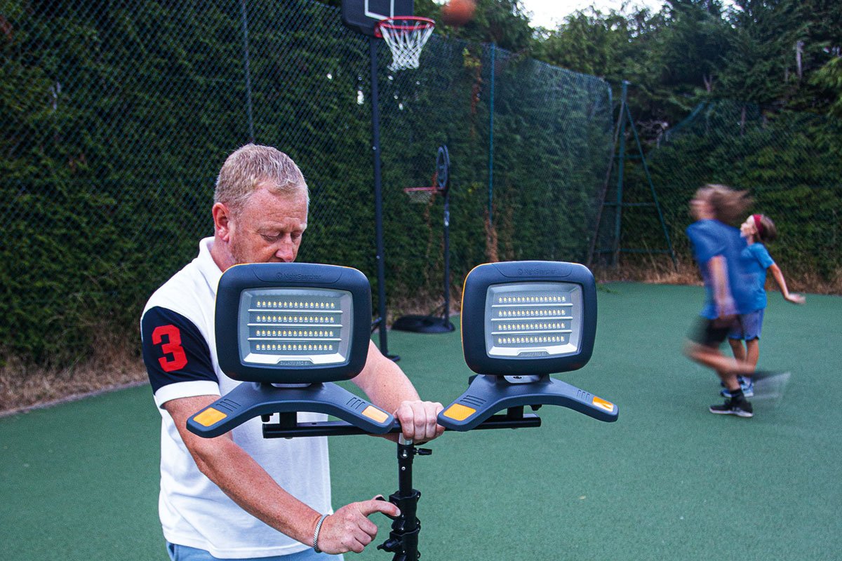 A man adjusts a Nightsearcher Galaxy Pro Twin Portable Floodlights unit on a stand at an outdoor basketball court while children play in the background, with trees and a basketball hoop visible behind them.