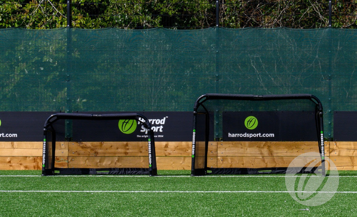 Two small black Finesse Folding Target Goals stand on artificial turf before a green mesh fence, trees in the background, with Finesse banners on the fence.
