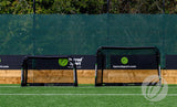 Two small black Finesse Folding Target Goals stand on artificial turf before a green mesh fence, trees in the background, with Finesse banners on the fence.