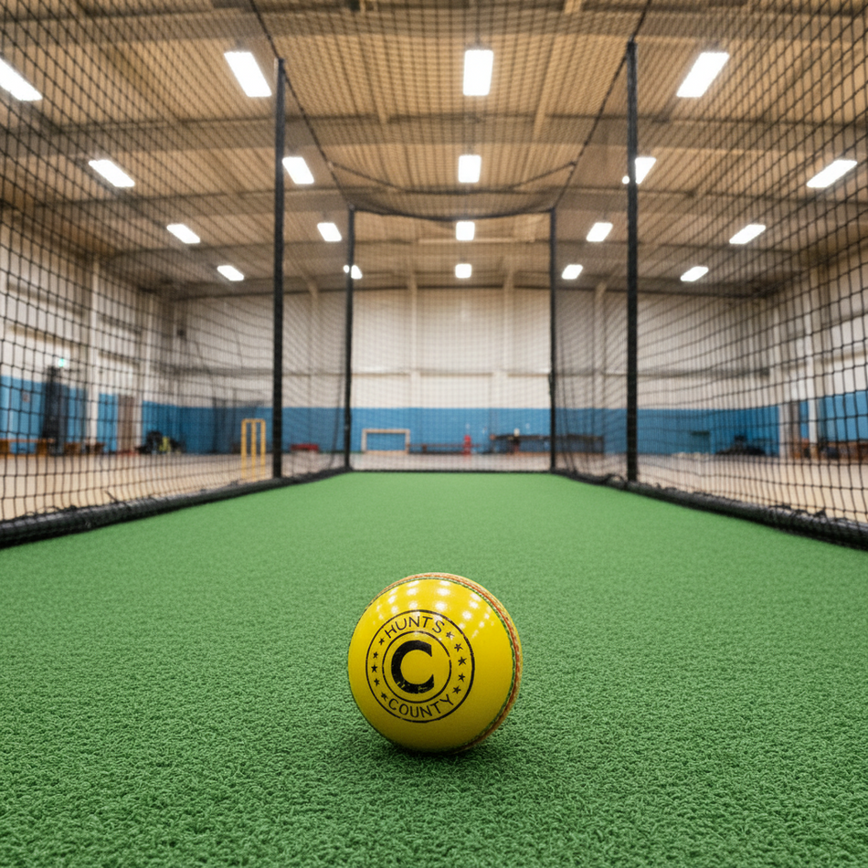 A yellow Hunts County Indoor Cricket Ball by Hunts County rests on green turf in an indoor practice net, with netting and sports hall visible in the background.