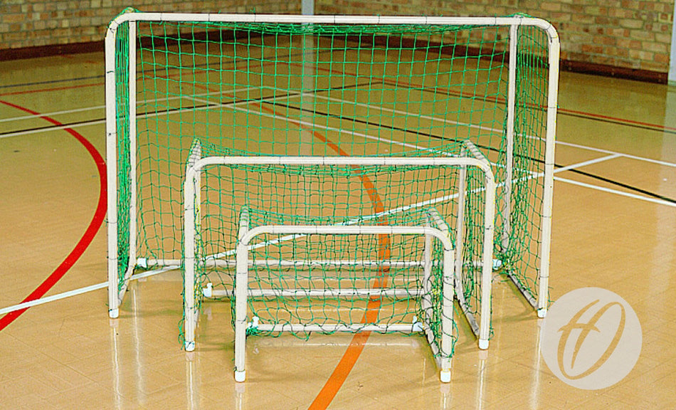 Three Harrod Sport Mini Hoc Goals (600mm x 900mm), white with green nets and folding frames, are nested together on an indoor wooden sports court with a brick wall—ideal for indoor use.