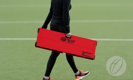 Wearing black athletic clothing, a person carries a folded Harrod Sport Folding Hockey Target Board across a green sports field.
