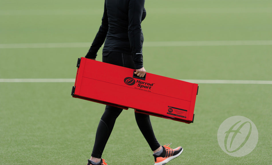 Wearing black athletic clothing, a person carries a folded Harrod Sport Folding Hockey Target Board across a green sports field.
