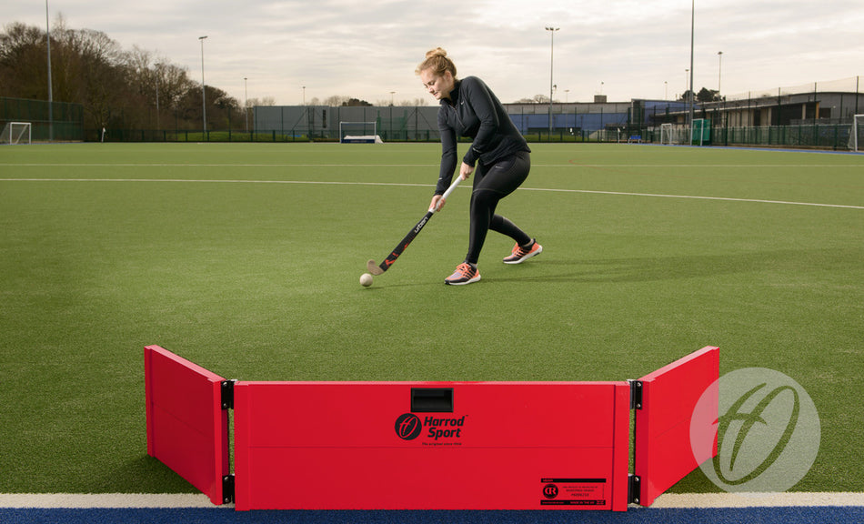 A person in sportswear plays field hockey on artificial turf, striking a ball with a hockey stick near a red Harrod Sport Folding Hockey Target Board. Sports facilities and trees are visible in the background.