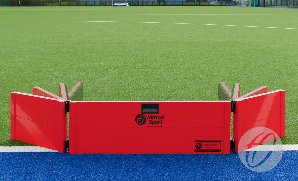 The Harrod Sport Folding Hockey Target Board, in bright red with adjustable friction hinges and foldable side panels, sits on green artificial turf by a blue and white boundary line—an ideal training aid for hockey players.