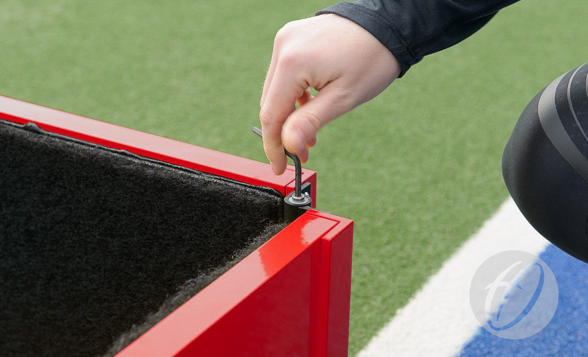 Using a hex key, a person tightens a bolt on the Harrod Sport Folding Hockey Target Board, which features adjustable friction hinges. The red metal training aid is set on artificial green turf near a blue and white surface.