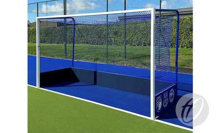 A Harrod Sport Match Hockey Goal Set – Blue with netting stands on blue artificial turf bordered by green, with a hedge in the background on a sunny day.