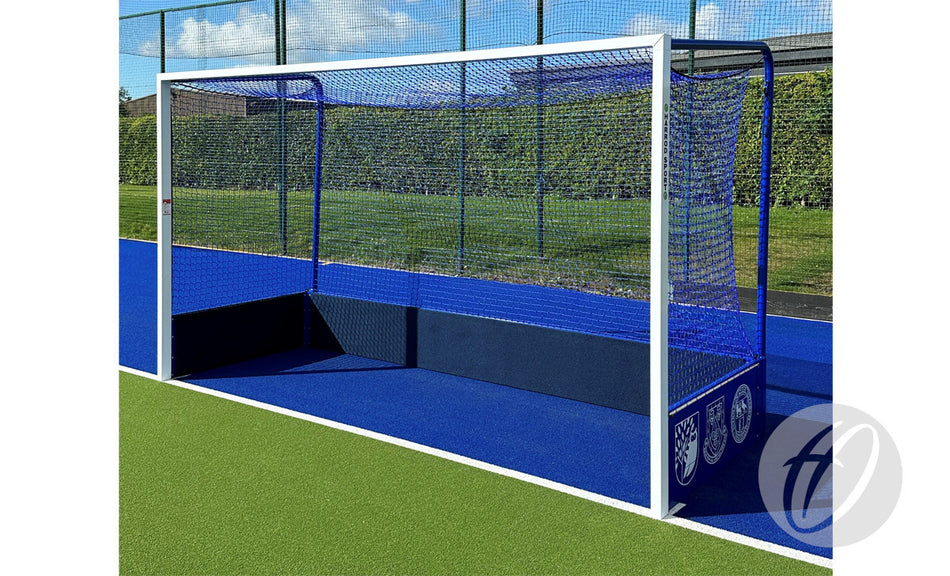 A Harrod Sport Match Hockey Goal Set – Blue with netting stands on blue artificial turf bordered by green, with a hedge in the background on a sunny day.
