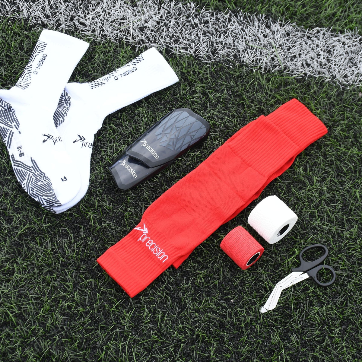 Red and white Precision cohesive tape rolls with football socks, shin pad and scissors on turf