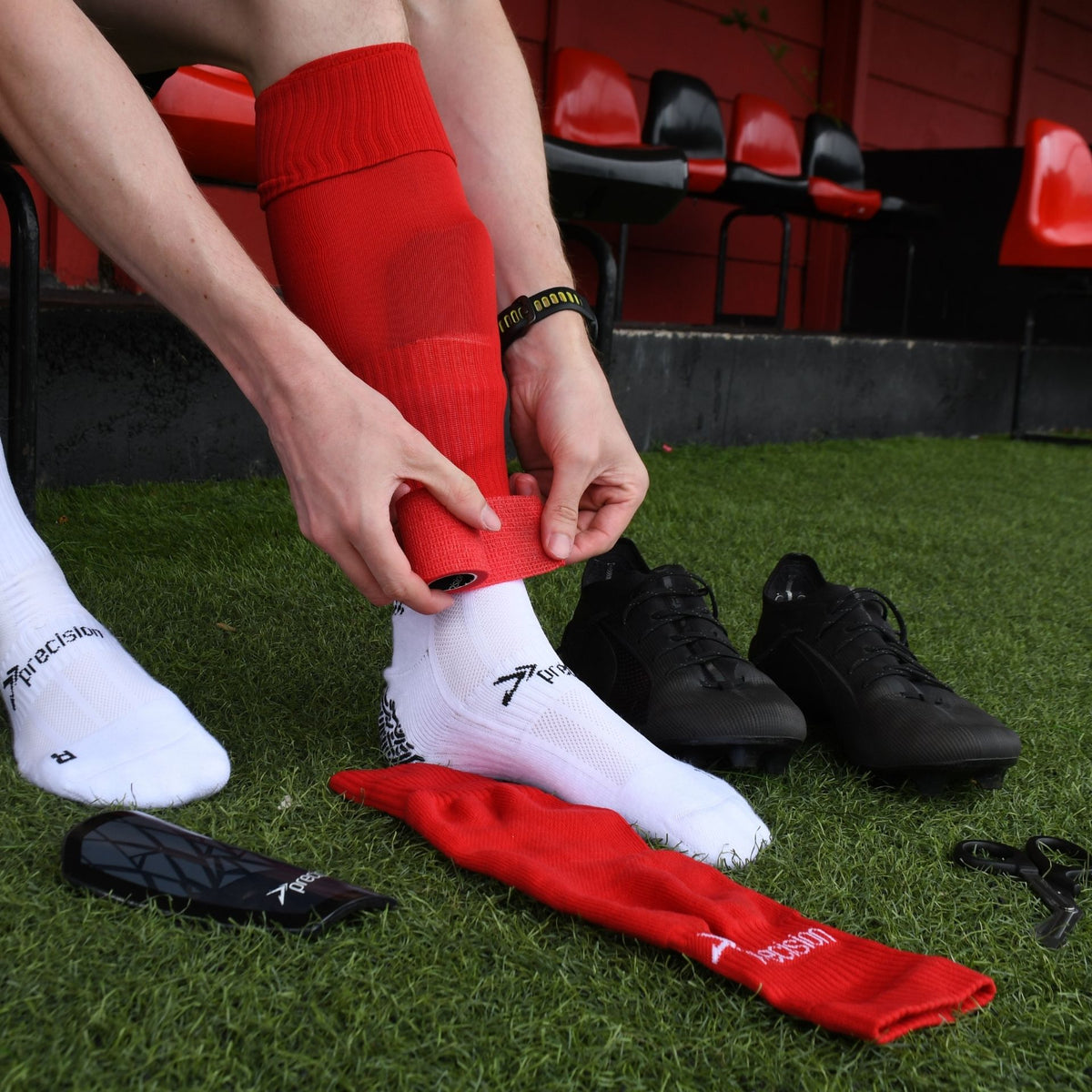 Player applying red Precision cohesive tape to a red sock pitch-side, stadium seats behind