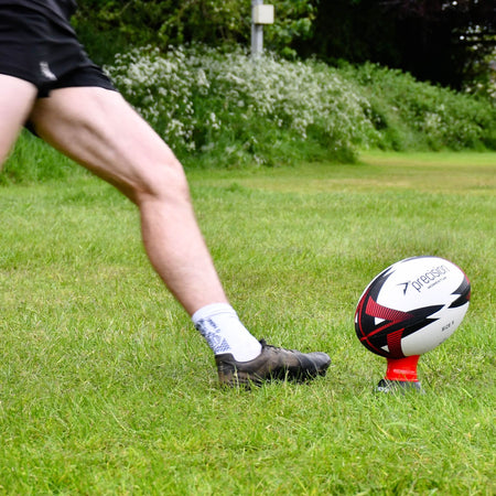 Player's leg swinging toward the red Precision Momentum Rugby Training Ball on a kicking tee