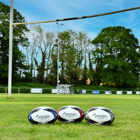 Blue, grey and red Precision Momentum Rugby Training Balls lined up on grass before rugby posts
